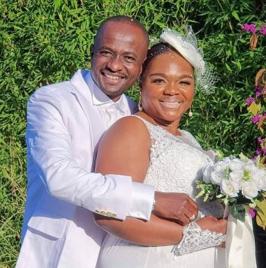 Un couple souriant en tenue de mariage, posant devant des fleurs et de la verdure.