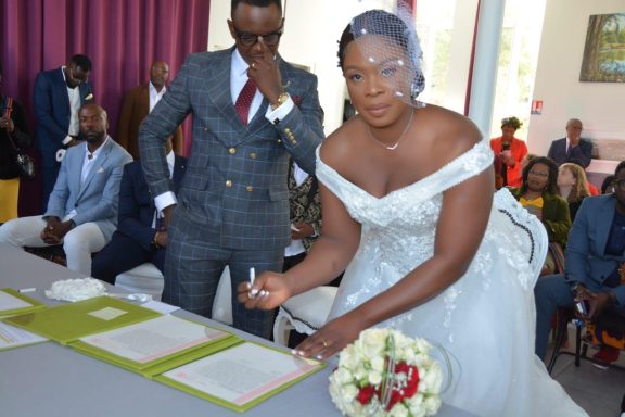 Une mariée signe des documents, entourée de ses invités. Un homme en arrière-plan la regarde.