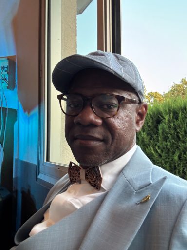 Homme souriant portant un costume clair et un nœud papillon, assis près d'une fenêtre.